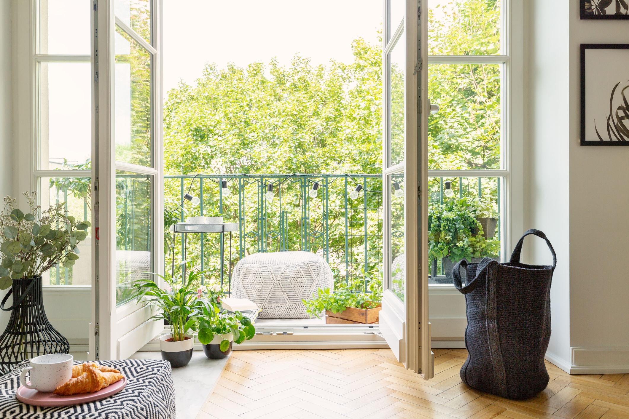 Open glass door from a living room interior into a city garden on a sunny balcony with green plants and comfy furniture